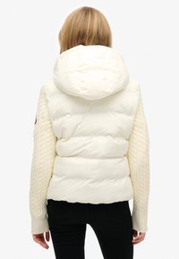 Kurtka puchowa w kolorze kremowym z kapturem, z rękawami w warkoczowym splocie i pikowanymi detalami, zaprojektowana dla ciepła i komfortu.