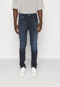 Hombre vestido con jeans ajustados de color azul oscuro, una camiseta de rayas gris claro y blanca de manga corta, y zapatillas blancas, apoyado contra un fondo liso.