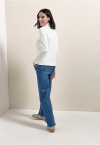 Valkoinen pitkähiha, rento malli, yhdistettynä sinisiin leveisiin farkkuihin. Yhdistetty vaaleanruskeisiin kenkiin, yksinkertainen tausta, kohde kääntynyt pois.