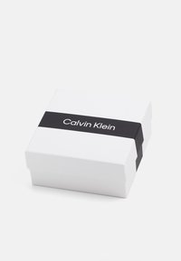 Caja de regalo de cartón blanco con una banda negra etiquetada "Calvin Klein" centrada en la parte frontal. De forma cuadrada con una tapa que se abre.