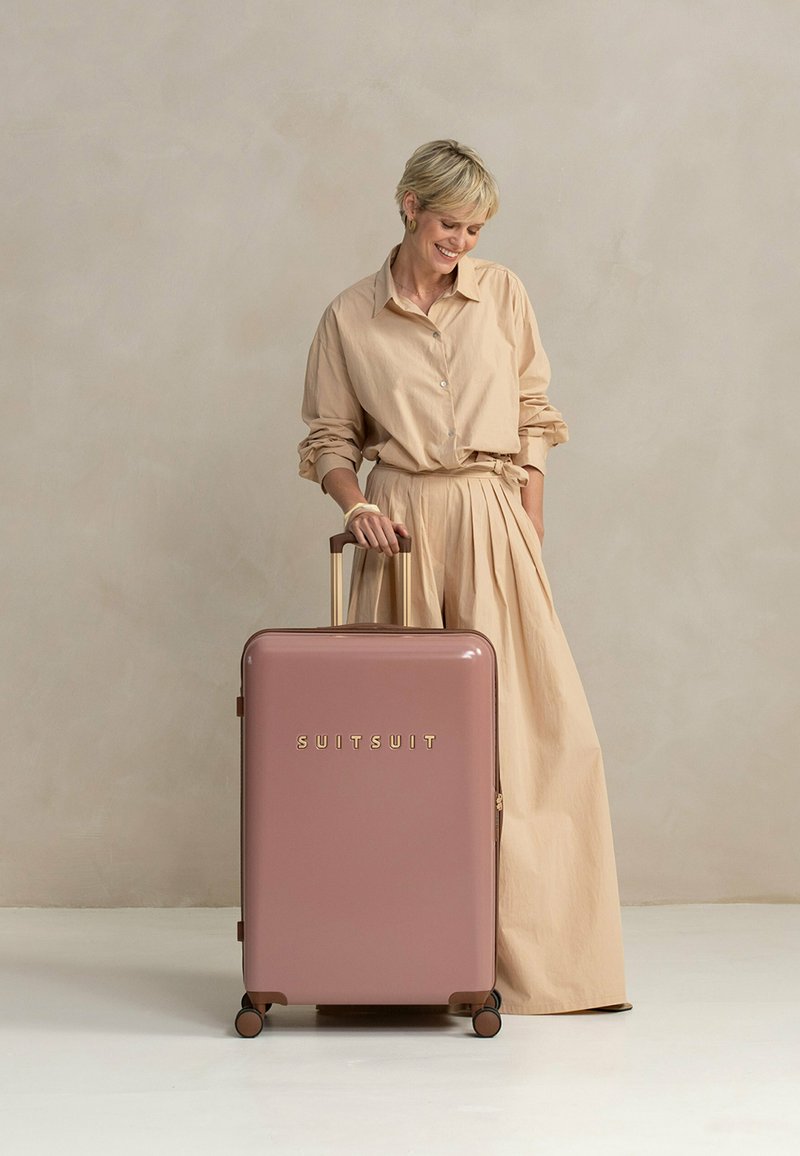 Vrouw in een beige outfit staat met haar hand op de handgreep van een roze koffer met wielen, gelabeld "SUITSUIT", tegen een effen achtergrond.