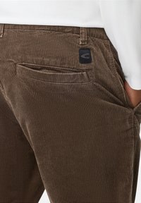 Brune kordfløjspants med et tekstureret ribmønster, sidelomme og en sort logo-patch bagpå. Skræddersyet silhuet.