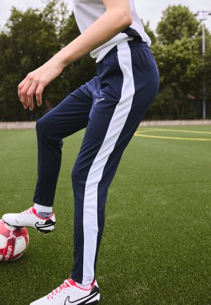 Pantalones deportivos de la marina con franjas laterales blancas, cinturilla elástica y logo. Llevados con zapatillas blancas y negras en hierba artificial.