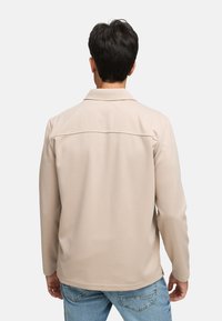 Chemise beige à manches longues avec un col, tissu texturé et une couture décorative sur la partie supérieure du dos, portée avec un jean bleu clair.