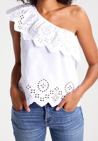White, one-shoulder top featuring a ruffled neckline, laser-cut detailing at the hem, and intricate patterns along the bottom edge.