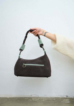 Ręka trzyma ciemnobrązową zamszową torebkę z plecionym uchwytem i jasnozielonym suwakiem na kieszeni na tle białej, gładkiej ściany.