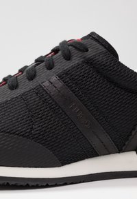 Chaussure de sport noire avec une tige en tissu texturé, des accents noirs mats, des œillets rouges et une semelle blanche. Présente le logo "HUGO" sur le côté.