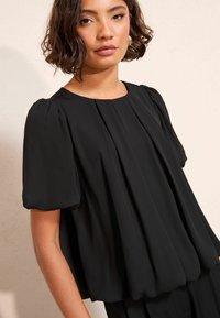 Fekete blúz puffos ujjakkal, redős kialakítással és kerek nyakkivágással. Puha, folyékony anyagból készült, sima textúrával.