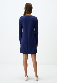 Abito blu navy a maniche lunghe con taglio dritto, dettagli della cucitura sul retro e scollo rotondo. Indossato con sneakers bianche.