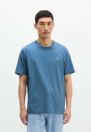 Homme debout portant un t-shirt bleu avec le logo MOP et un jean bleu clair sur un fond blanc uni.