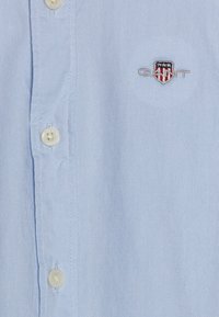 Camicia button-up azzurro chiaro realizzata in tessuto testurizzato, con un piccolo logo ricamato con accenti rossi, bianchi e blu sul petto.