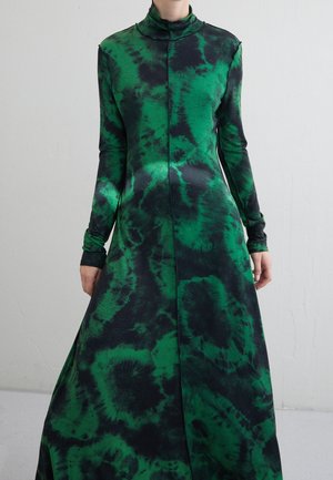 Długa sukienka maxi z długim rękawem i wysokim kołnierzem, w zielono-czarny wzór tie-dye, noszona przez stojącą osobę na jednolitym tle.
