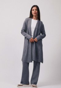 Une femme porte un long cardigan gris, un pantalon large assorti, une chemise blanche et des baskets blanches, posant devant un fond uni.