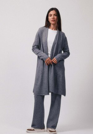 Une femme porte un long cardigan gris, un pantalon large assorti, une chemise blanche et des baskets blanches, posant devant un fond uni.