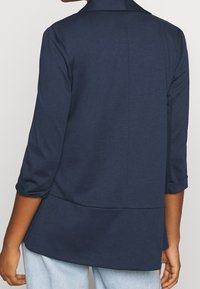 Mörkblå, lätt jerseyblazer med trekvartslånga ärmar, skräddarsydd passform och hög krage, med en mjuk textur och subtila sömmar.