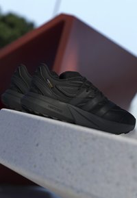 Sorte atletikskøjter med tykke såler og Cordura-stoflabel, placeret på en hvid overflade med en rød geometrisk baggrund.
