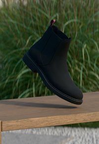 Schwarzer Knöchelstiefel mit schlankem Design, glattem synthetischem Material, runder Zehenform und einem roten und weißen Zugband an der Ferse.