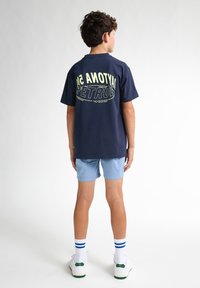 Camiseta de algodón azul marino con gran texto gráfico amarillo-verde en la parte posterior. Combinada con pantalones cortos azul claro y zapatillas blancas con acentos verdes.