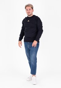 Czarna bluza z okrągłym dekoltem, złoty detal z logo, w połączeniu z niebieskimi dżinsami i białymi sneakersami. Tkanina wydaje się miękka i swobodna.