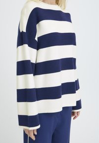 Mujer vistiendo un suéter de rayas horizontales anchas en azul marino y blanco, con mangas largas y pantalones azul marino.