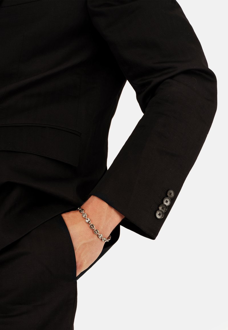 Eine Person trägt einen schwarzen Anzug und hat ihre rechte Hand in der Tasche, wobei ein silbernes Armband an ihrem Handgelenk zu sehen ist.