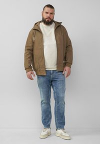 Bruine hoodie-jas met een rits aan de voorkant, gedragen over een licht gebreid sweater, gecombineerd met blauwe jeans en witte sneakers met beige accenten.