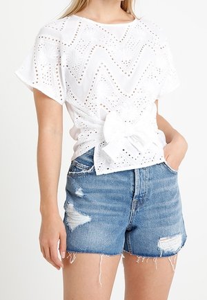 Hvid blondebluse med korte flagrende ærmer og sløjfe foran, kombineret med slidte blå denimshorts.