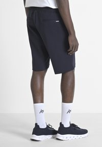 Pantaloni sportivi blu navy con vita regolabile tramite cordino, dotati di due tasche posteriori e una texture liscia. Indossati con calzini bianchi e sneakers blu navy.