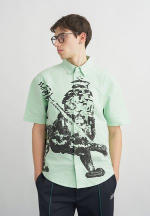 Nuori mies, jolla on silmälasit ja päällään vaaleanvihreä nappipaita, jossa on suuri musta graafinen kuvio, sekä tummat housut ja kädet taskuissa.