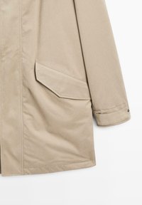Beige vattentät jacka med rak skärning, långa ärmar, en stor sned ficka på vänster sida och sydda ytterkanter.