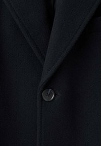 Cappotto di lana nera con revers a punta e chiusura con un solo bottone nero, con una finitura morbida e strutturata.