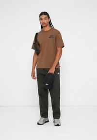 Rudas Nike marškinėliai su juodu logotipu, žali lauko kelnės su kišenėmis, pilki sportbačiai ir juoda kepurė laikoma rankoje prieš baltą fono.