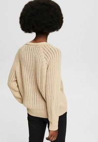 Beiger gestrickter Pullover mit Lochmuster, gerippten Bündchen und Saum. Der Rücken ist mit strukturiertem Strickmuster versehen und hat eine lockere Passform. Kombiniert mit schwarzen Hosen.