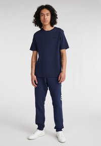 Camiseta de algodón navy con cuello redondo y mangas cortas, combinada con joggers a juego que presentan texto de logo en blanco; usada con zapatillas blancas.