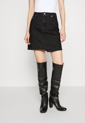 Femme portant une mini-jupe en denim noir avec une fente à l'avant, associée à des bottes en cuir noir montantes à talons et un haut blanc sans manches.