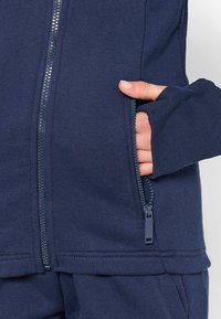 Námornícky zip-up mikina s hladkou textúrou, obsahujúca predný zips a bočnú kapsu. Otvory na palce v rukávoch. Jednoduchý, funkčný dizajn.