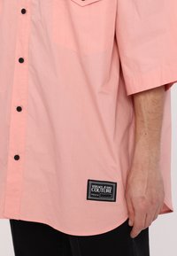 Camisa oversize de color rosa claro, confeccionada en un tejido suave, con tapeta de botones, mangas cortas y una etiqueta negra que dice "VERSACE JEANS COUTURE".