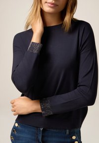Vrouw in een marineblauwe top met lange mouwen en versierde manchetten, en blauwe jeans met gouden knopen, poseert met één hand bij haar nek.