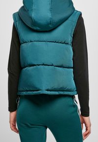 Tyrkysová péřová vesta s kapucí, má prošívaný design a elastické stahovací šňůrky na spodním lemu. Nosit přes černé tričko s dlouhým rukávem.