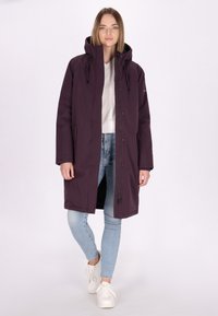 Langer lila Mantel mit Kapuze, Reißverschluss vorne und Druckknopfverschluss, ausgestattet mit seitlichen Taschen und einem lockeren Schnitt, getragen über einem weißen Hemd und blauen Jeans.