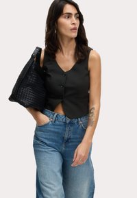 Gilet scuro corto con quattro bottoni e scollo a V, abbinato a jeans larghi di colore azzurro chiaro. Una borsa nera testurizzata è indossata a tracolla.