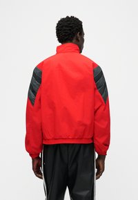 Άνδρας με κοντά σγουρά μαλλιά, φοράει κόκκινο μπουφάν με μαύρα σχέδια σε σχήμα βέλους στα μανίκια και μαύρο παντελόνι με λευκές ρίγες, πίσω όψη.