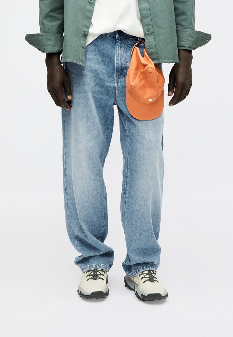 Man wearing light blue jeans, white sneakers, green shirt, with orange cap hanging from jeans pocket.