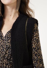 Gilet en fourrure noire texturée avec un large col, associé à un haut à motifs noir et doré, doté d'un décolleté en V et d'une taille élastique.