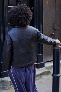 Osoba s kučeravými vlasmi, oblečená v čiernej koženej bunde a modrých nohaviciach, sa opiera o čierny stĺp pri tmavých dverách s číslom 18.