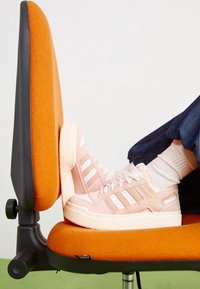 Persikofärgade mockasneakers med vita ränder på sidorna, rund tå och tjock krämfärgad sula, vilande på en orange kontorsstol.