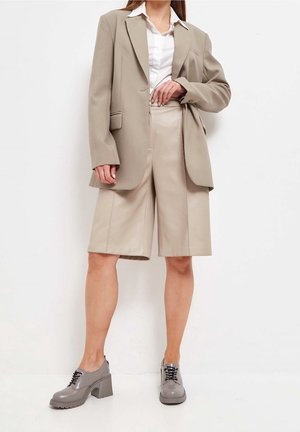 Femme portant un blazer beige, un short assorti à mi-cuisses, une chemise blanche et des chaussures à talons épais grises à lacets, se tenant devant un fond blanc.