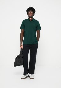 Camisa polo verde, com mangas curtas, logo bordado. Calças largas pretas, mala acolchoada preta, ténis brancos com sola castanha.