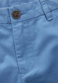 Tessuto di cotone azzurro chiaro con una consistenza liscia; presenta un bottone rotondo marrone con incisioni e cuciture visibili lungo le cuciture.