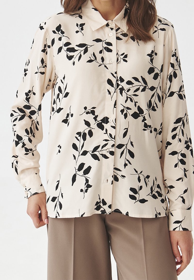 Femme portant une chemise beige à manches longues boutonnée avec un motif de branche feuillue noire, associée à un pantalon marron clair.